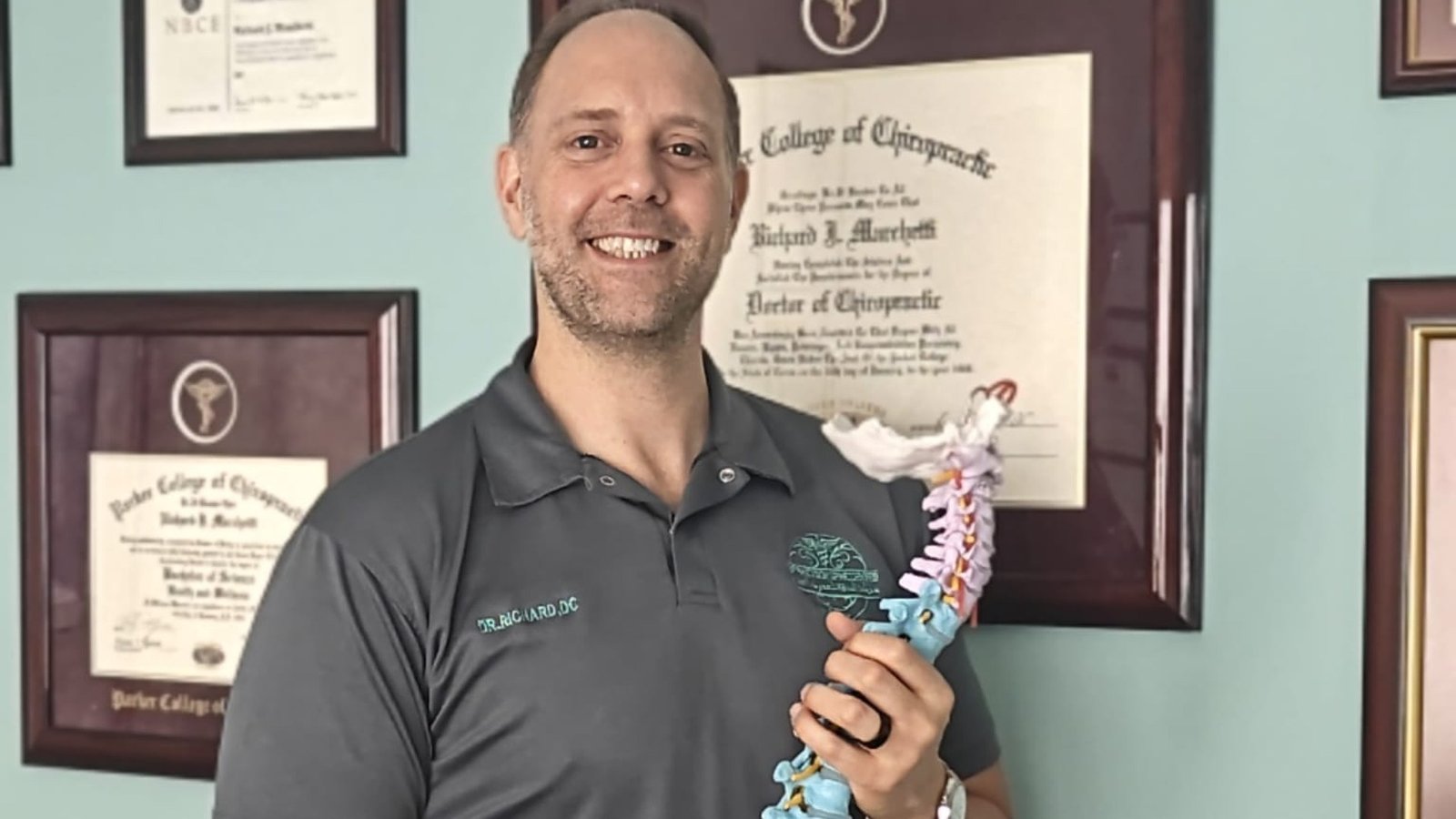Dr. Richard Marchetti at CBP Precision Spine Center in Muscat, holding an anatomical spine model in front of his framed chiropractic diplomas and certifications