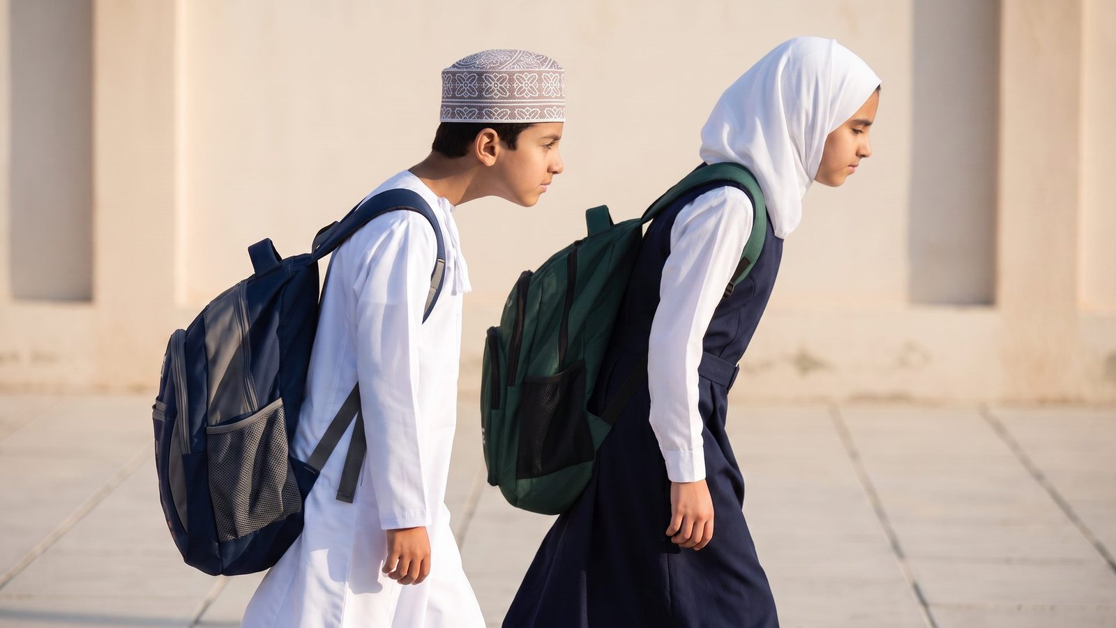 طفلان عُمانيان يحملان حقيبتَي ظهرٍ ثقيلتين ويميلان إلى الأمام مع تقدّم الرأس عن الكتفين